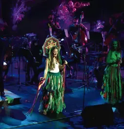 Cantante luciendo tocado artesanal y falda de tiras verdes actuando junto a músicos en vivo.