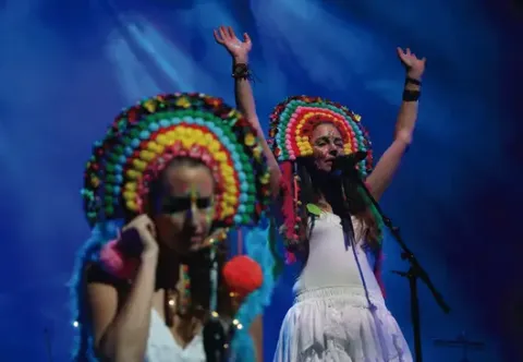 Artistas con vistosos tocados de pompones y vestidos blancos cantando con los brazos alzados en el escenario.