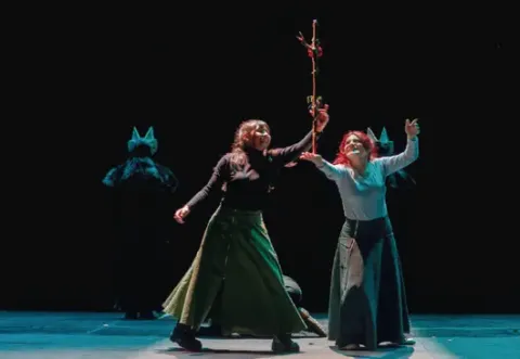 Actrices sonrientes en un escenario con una rama floral, figuras con máscaras de animales al fondo.