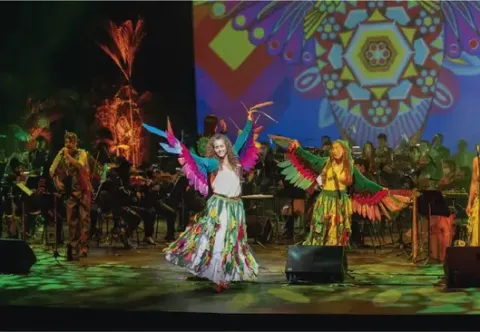 Bailarinas vistiendo alas de colores y faldas estampadas danzando en un escenario musical festivo.