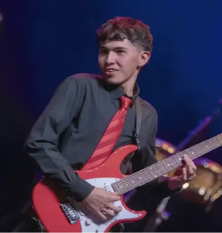 Joven músico con camisa oscura y corbata roja tocando una guitarra eléctrica en el escenario.