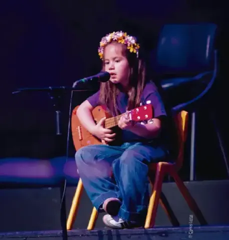 Niña pequeña cantando frente al micrófono mientras toca una pequeña guitarra.