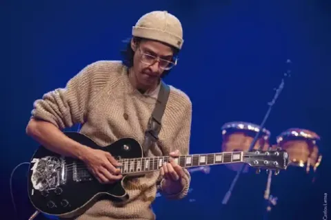 Músico tocando concentrado una guitarra eléctrica negra en el escenario.