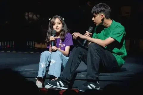 Dos jóvenes sentados en el borde del escenario conversando con micrófonos en mano.