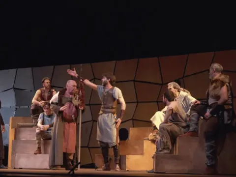 Elenco teatral con vestuario de época clásica actuando sobre una estructura de gradas en el escenario.