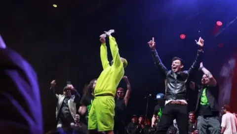Celebración en escenario: Hombre de neón levanta objeto, otro salta con brazos arriba.