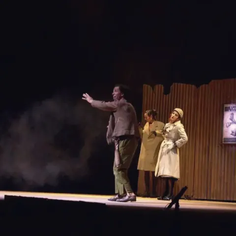 Actores en escena: hombre gesticulando, dos mujeres observan, con niebla y un póster de gramófono.
