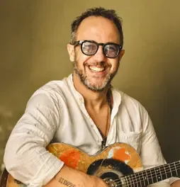 César López sonriendo con gafas de marco negro mientras sostiene una guitarra acústica.