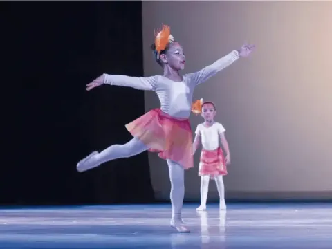 Dos niñas con vestuario y adornos de plumas ejecutando una danza en el escenario.