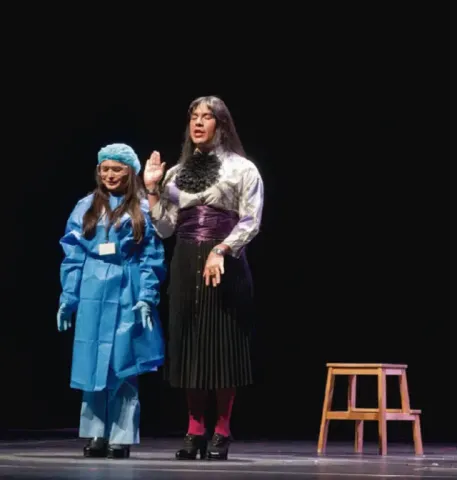 Dos actores representando una escena cómica o dramática junto a un taburete de madera.
