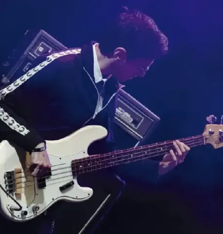 Músico de perfil tocando un bajo eléctrico blanco en un escenario iluminado con luz azul.