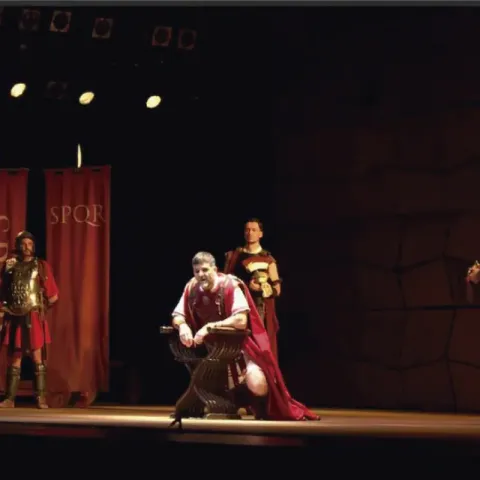 Actores en escena con vestuario romano (uno sentado), estandartes SPQR y luces.