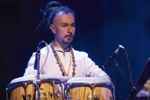 Percusionista con rastas y vestimenta clara tocando las congas en vivo.