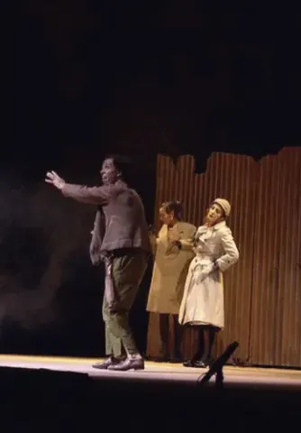Tres actores interpretando una escena dramática frente a una pared de láminas de madera.