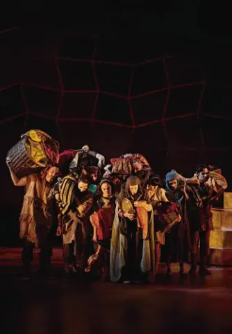 Grupo de actores caracterizados cargando cestas y bultos en una escena dramática sobre el escenario.