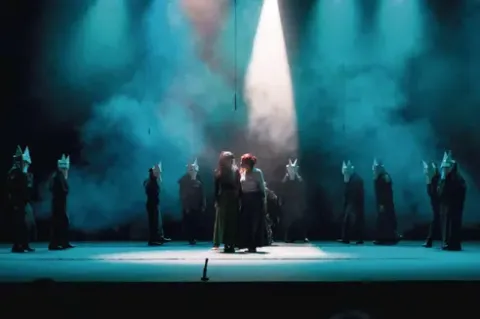 Dos actrices en el centro del escenario bajo un reflector rodadas por figuras enmascaradas.