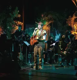 Músico tocando la guitarra acústica y cantando en el centro del escenario con la orquesta de fondo.