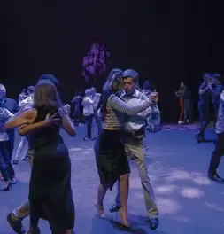 Parejas abrazadas bailando milonga bajo la iluminación tenue del escenario.