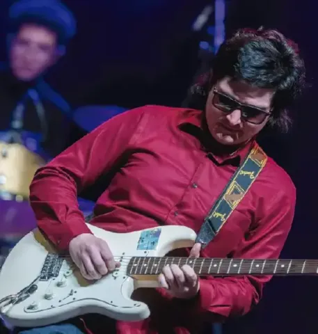 Guitarrista con gafas de sol y camisa roja ejecutando un solo en su guitarra eléctrica blanca.