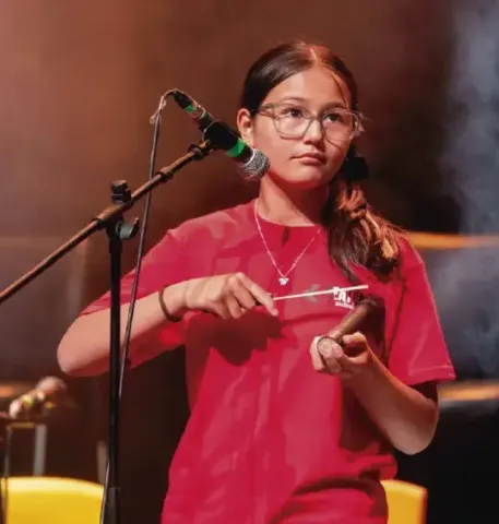 Joven con gafas tocando un instrumento de percusión menor frente a un micrófono.