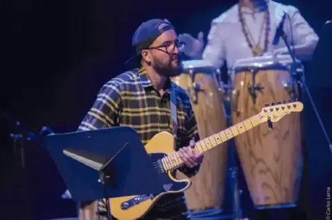 Guitarrista con gorra y camisa a cuadros tocando una guitarra eléctrica frente a su atril.