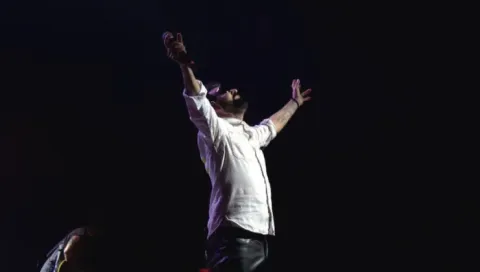 Cantante con camisa blanca y brazos levantados sosteniendo un micrófono bajo la iluminación del escenario.