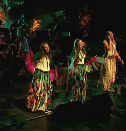 Tres cantantes vestidas con atuendos folclóricos y alas vistosas interpretando una canción frente al público.
