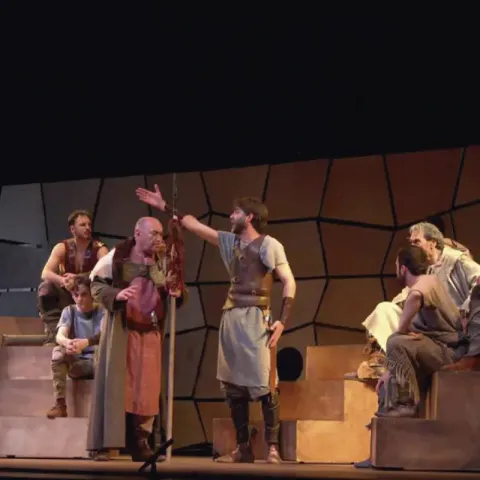 Actores en un escenario teatral. Un hombre gesticula con el brazo levantado, otros lo observan.