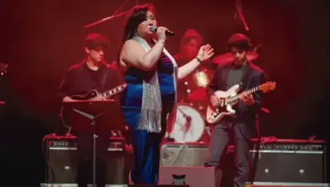Cantante en vestido azul interpretando un tema junto a su banda en vivo.