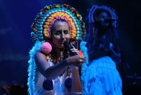 Artista con tocado colorido de pompones y maquillaje brillante cantando frente a un micrófono en el escenario.