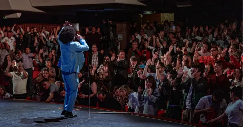 Artista en traje azul animando al público desde el escenario en un teatro lleno.