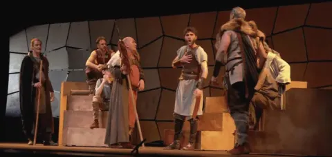 Elenco de teatro clásico interpretando una escena dramática sobre un escenario con gradas.